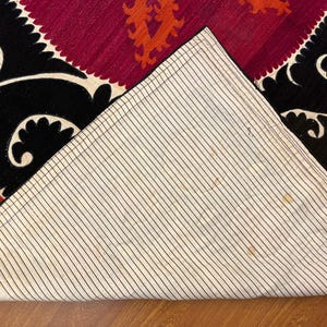May include: Close-up of a section of a rug with a geometric design. The rug features a black border with white scrollwork, a burgundy center with orange accents, and a striped off-white backing.