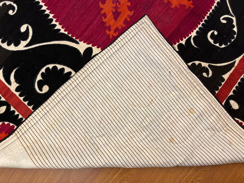 May include: Close-up of a section of a rug with a geometric design. The rug features a black border with white scrollwork, a burgundy center with orange accents, and a striped off-white backing.