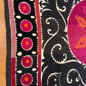 May include: Close-up of a section of a textile with a black background and white, red, and pink floral and scroll designs. The textile has a border with a black edge and a pink and white striped section.