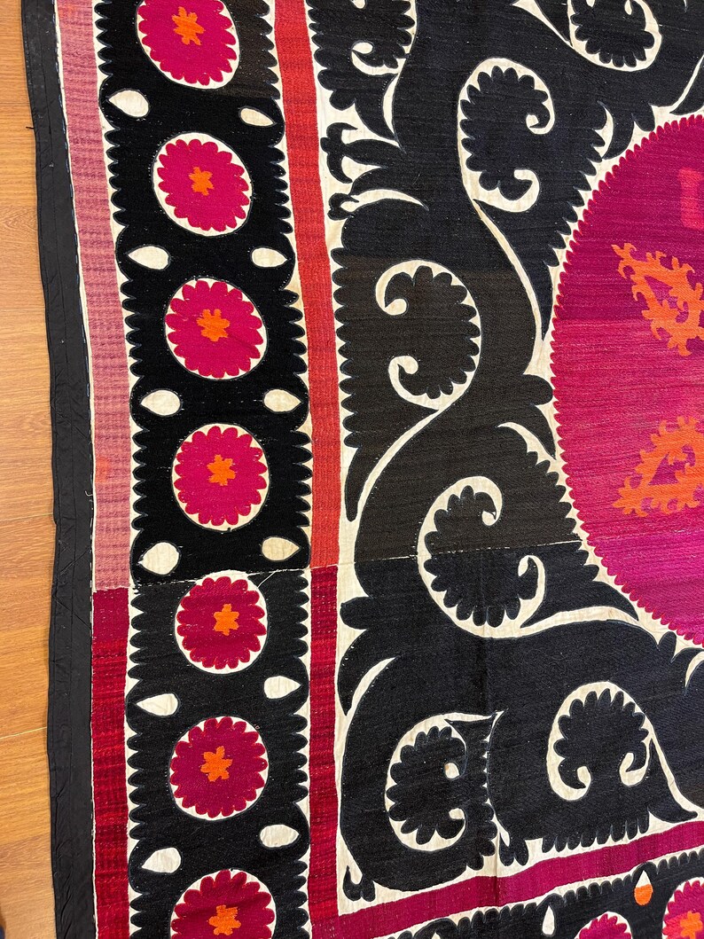 May include: Close-up of a section of a textile with a black background and white, red, and pink floral and scroll designs. The textile has a border with a black edge and a pink and white striped section.