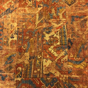May include: Close-up of a vintage rug with a patchwork design. The rug features a complex pattern in shades of red, brown, blue, and beige. The texture appears woven, with visible threads and a slightly worn appearance, suggesting age and use.