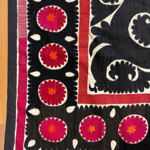 May include: Close-up of a textile with a black background featuring repeating floral patterns in red and orange. The design includes white, scalloped edges and a red border. The textile has a black border.