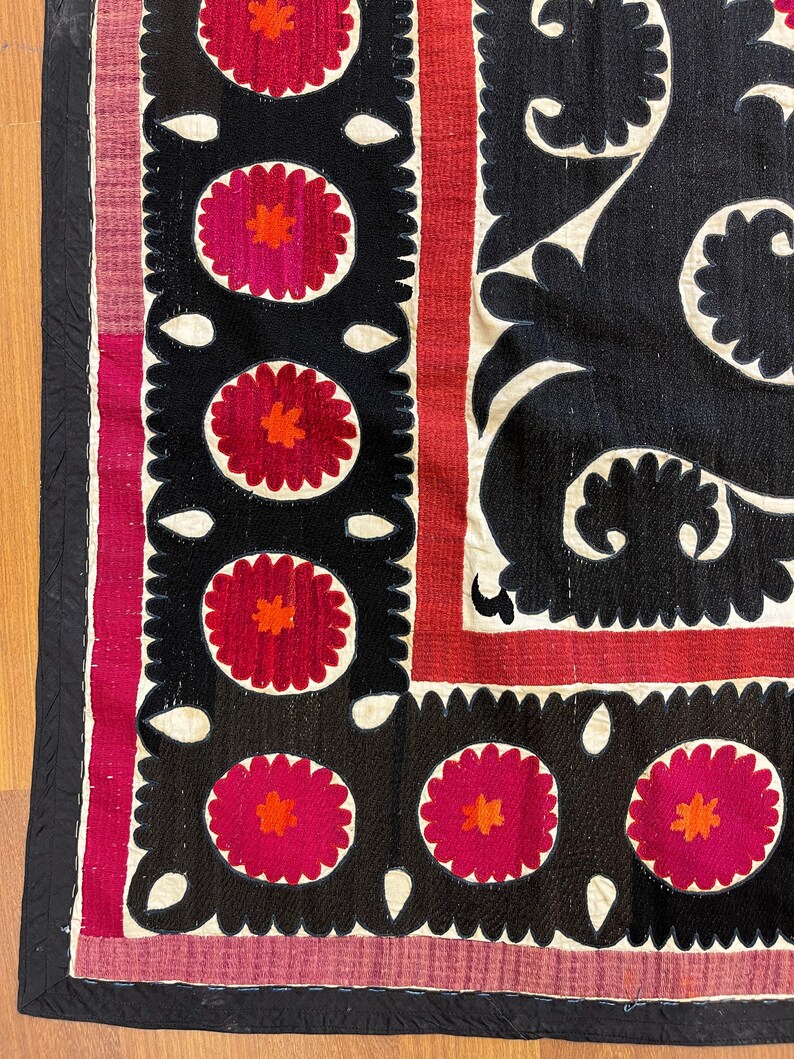 May include: Close-up of a textile with a black background featuring repeating floral patterns in red and orange. The design includes white, scalloped edges and a red border. The textile has a black border.