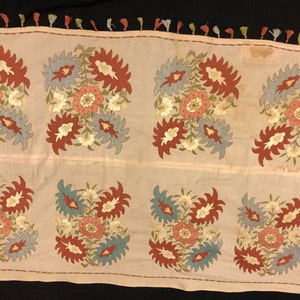 May include: A rectangular embroidered fabric panel with a floral design. The panel is made of a light pink fabric and features a repeating pattern of blue, red, and white flowers.