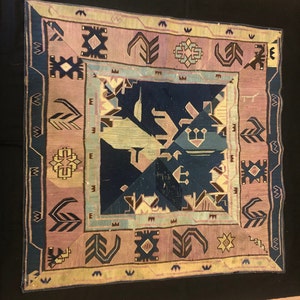 Op de afbeelding: Een vintage tapijt met een geometrisch patroon in tinten roze, blauw en beige. Het tapijt heeft een centraal vierkant met een complex ontwerp en een rand met herhalende motieven.
