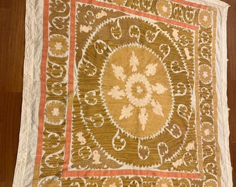May include: A square, decorative textile with a central floral design in shades of gold and cream, surrounded by a border of peach and brown. The textile has a white border and appears to be made of a soft, possibly quilted material.