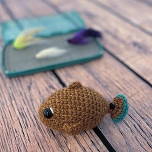 May include: A brown crocheted fish with a teal tail fin and a black eye. The fish is sitting on a wooden surface.