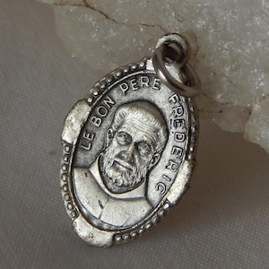 May include: An oval silver pendant with the inscription "LE BON PERE FREDERIC" around a portrait of a bearded man. The pendant has a loop for attaching to a chain. The metal has an aged look, with detailed design.