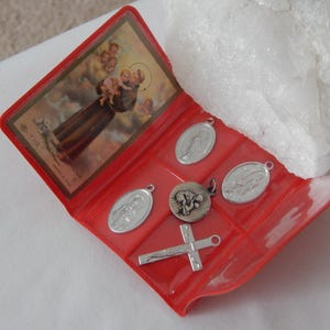 Könnte beinhalten: Eine rote Plastikbox mit mehreren religiösen Anhängern und einem kleinen Bild. Die Anhänger sind silberfarben und umfassen ein Kreuz, ovale Medaillons und einen runden Anhänger. Das Bild zeigt eine religiöse Figur.