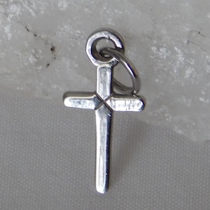 Könnte beinhalten: Ein kleiner, silberfarbener Kreuzanhänger mit einer Schlaufe oben zum Befestigen an einer Kette. Das Kreuz hat ein schlichtes, klassisches Design mit leicht strukturierter Oberfläche. Der Anhänger wird vor einem hellen Hintergrund gezeigt.