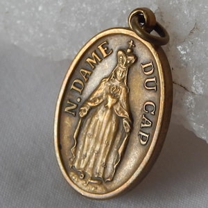 May include: An antique gold-coloured oval pendant, inscribed with "N. DAME DU CAP" and featuring a detailed image of a robed figure. The pendant has a loop for attachment and shows signs of age.