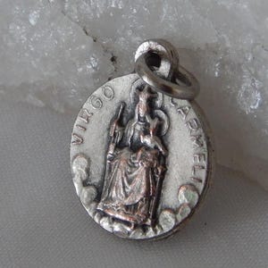 May include: A silver-toned pendant with the words "VIRGO CARMELI" encircling a detailed image of a robed figure. The pendant has a small loop at the top for attaching to a chain or bracelet. The background is a light, textured surface.
