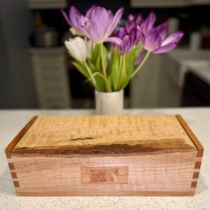 May include: A handcrafted wooden box with a light-colored, textured top and darker wood trim. The box features dovetail joints and a decorative inlay. A vase of purple and white tulips sits in the background.