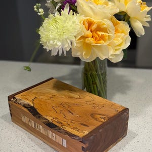 May include: A rectangular wooden box featuring a natural live-edge top with dark grain patterns, contrasting dark walnut sides, and a front panel accented with a vertical inlay of light and dark wood strips joined with visible finger joint corners.