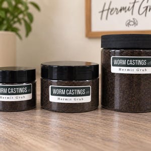 May include: Three clear cylindrical plastic jars of varying sizes filled with dark brown worm castings. Each jar features a black screw-top lid and a white label with black text reading WORM CASTINGS and Hermit Grub with a small crab icon.