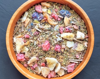 May include: A round wooden bowl filled with a loose-leaf tea blend containing dried green herbs, small brown twigs, bright red strawberry pieces, yellow pineapple chunks, and dark blue dried berries, viewed from directly above.