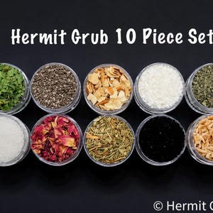 May include: A ten-piece set of hermit crab food, each in a clear plastic container. The set includes various dried ingredients like green herbs, seeds, and dried fruits, arranged on a black surface. Text on the image reads "Hermit Grub 10 Piece Set".