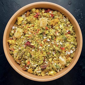 Op de afbeelding: Een ronde houten kom gevuld met een droge kruidentheemix met gele, groene en rode gedroogde ingrediënten. Het mengsel bevat kleine stukjes gedroogd fruit, zaden, kruiden en grotere, doorschijnende gele stukjes gekristalliseerde suiker.