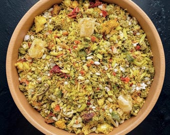 May include: A round wooden bowl filled with a dry herbal tea blend containing yellow, green, and red dried ingredients. The mixture includes small pieces of dried fruit, seeds, herbs, and larger translucent yellow chunks of crystallized sugar.