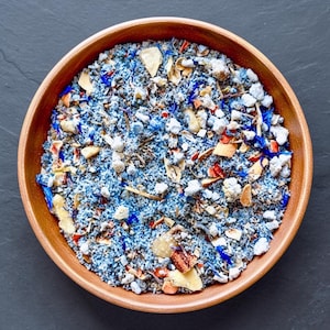 May include: A round wooden bowl filled with a textured mixture of blue-dyed coarse grains, small white mineral-like chunks, dried orange peel fragments, and vibrant blue cornflower petals, creating a multi-colored and granular composition.