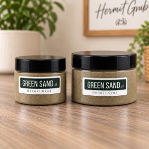 May include: Two clear plastic jars with black screw-on lids containing fine green sand. Each jar features a white rectangular label with dark green text that reads GREEN SAND and Hermit Grub, accompanied by a small line drawing of a crab.
