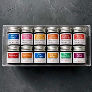 Op de afbeelding: Een doorzichtige acrylbak met twaalf kleine glazen potjes met zilveren deksels, gerangschikt in twee rijen van zes. Elk potje heeft een wit etiket met unieke tekst, waaronder Campfire Nights, Birthday Cake, All Star Breakfast, Tuna Casserole, Cranberry Pumpkin Spice, Flame Thrower, Jambalaya, Street Tacos, Blue Octopus, Tropical Delight, Mint Chocolate Chip en Tigers Blood.