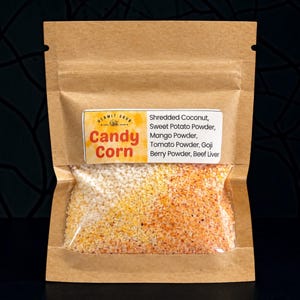 Op de afbeelding: Een bruine kraftpapieren zak met een doorzichtig venster met een korrelig mengsel van witte, gele en oranje deeltjes. Het etiket vermeldt Hermit Grub Candy Corn en ingrediënten zoals geraspte kokos, zoete aardappelpoeder, mangopoeder, tomatenpoeder, gojibessenpoeder en runderlever.
