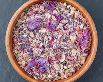 May include: A round wooden bowl filled with a dry herbal tea blend containing visible pieces of dried purple hibiscus flowers, light brown grains, small green herbal stems, and various fragmented botanical ingredients in shades of pink and tan.