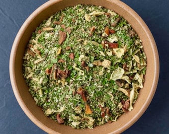 May include: A round, light brown ceramic bowl filled with a dry seasoning blend. The mixture consists of finely ground green herbs, small white granules, and scattered pieces of dried red and yellow vegetables, creating a textured appearance.