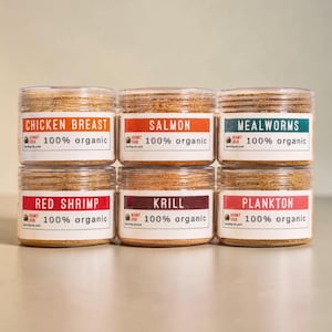 May include: Six clear plastic jars filled with organic food for hermit crabs, arranged in two rows. Each jar features a white label with the text 100% organic and a specific ingredient name: Chicken Breast, Salmon, Mealworms, Red Shrimp, Krill, or Plankton.