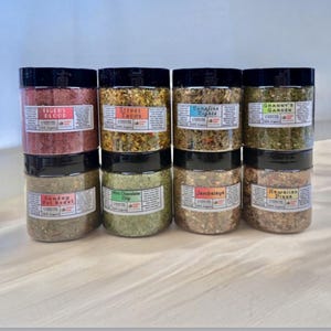 May include: Eight clear plastic jars with black lids, arranged in two rows of four. Each jar contains a different colored dry spice blend and features a white label with text including Tigers Blood, Street Tacos, Campfire Nights, Granny's Garden, Sunday Pot Roast, Mint Chocolate Chip, Jambalaya, and Hawaiian Pizza.