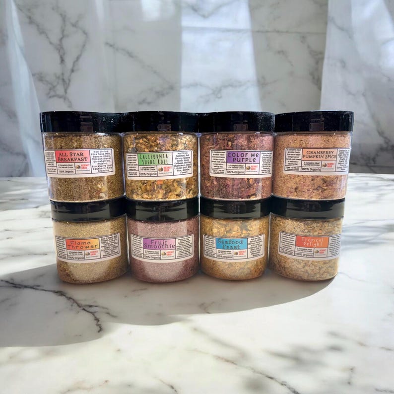 May include: Eight clear plastic jars with black lids arranged in two rows of four. Each jar contains a different colored organic food blend with a rectangular label featuring names like All Star Breakfast, California Sushi Roll, Color Me Purple, and Flame Thrower.