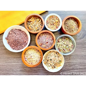 May include: Eight small bowls containing various dry food mixtures, including grains, herbs, and dried fruit pieces. The bowls are made of wood, white ceramic, or clear glass. The text "Hermit Grub 2022" is visible in the bottom right corner.
