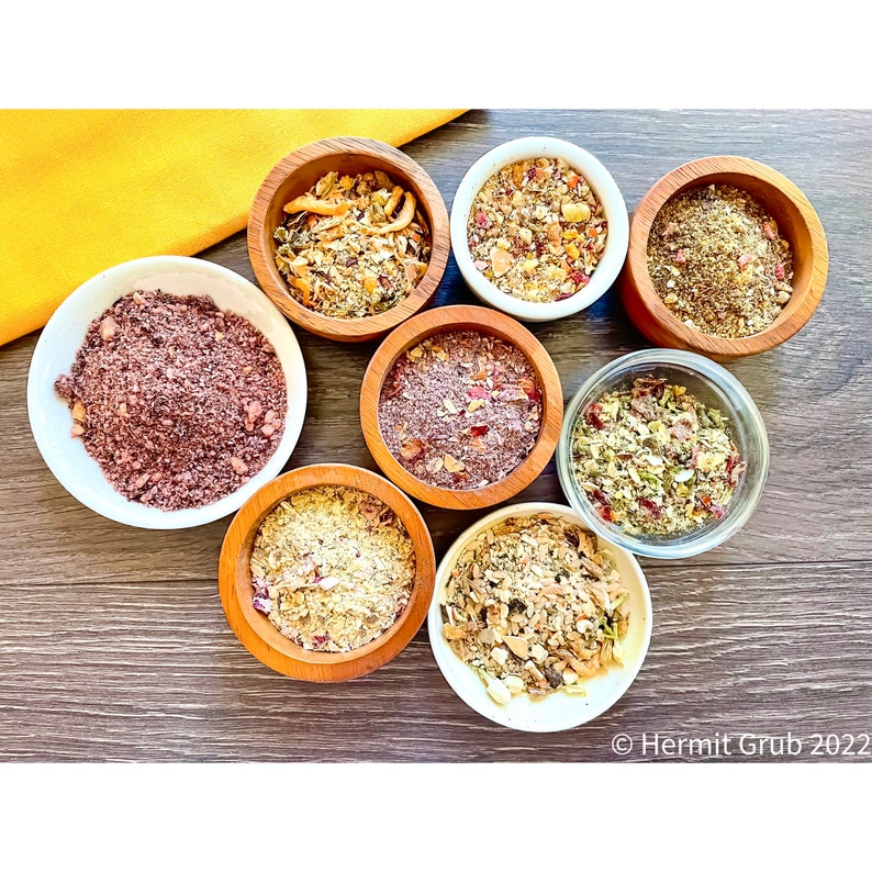 May include: Eight small bowls containing various dry food mixtures, including grains, herbs, and dried fruit pieces. The bowls are made of wood, white ceramic, or clear glass. The text "Hermit Grub 2022" is visible in the bottom right corner.