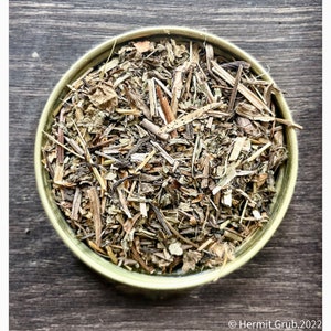 May include: A light green bowl filled with dried herbs and leaves. The herbs are a mix of green, brown, and tan, with visible stems and leaf fragments. The bowl sits on a dark wooden surface.