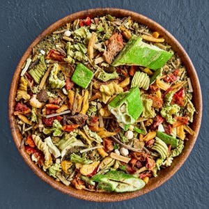 Pode incluir: Uma tigela de madeira redonda cheia de uma mistura seca de vegetais desidratados coloridos, incluindo fatias de quiabo verde brilhante, pedaços de tomate vermelho, grãos de milho amarelos e várias ervas e talos verdes fibrosos, vista de cima.