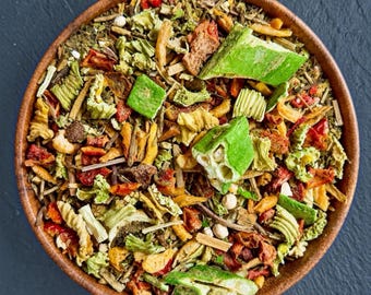 May include: A round wooden bowl filled with a dry mixture of colorful dehydrated vegetables, including bright green okra slices, red tomato pieces, yellow corn kernels, and various fibrous green herbs and stalks, viewed from directly above.