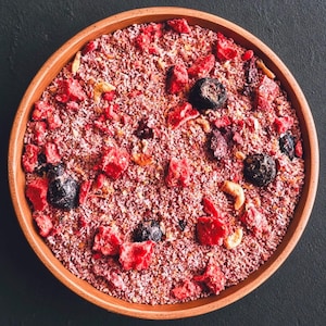 May include: A round terracotta bowl filled with a mixture of shredded coconut, freeze-dried red strawberries, dark blueberries, and light-colored nuts. The texture is granular and varied, featuring bright red, deep purple, and tan color tones.