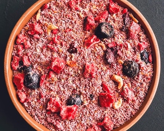 May include: A round terracotta bowl filled with a mixture of shredded coconut, freeze-dried red strawberries, dark blueberries, and light-colored nuts. The texture is granular and varied, featuring bright red, deep purple, and tan color tones.