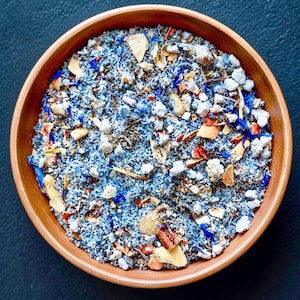 May include: A round wooden bowl filled with a loose-leaf tea blend. The mixture contains blue-tinted tea leaves, white rock-like pieces, dried orange peel fragments, and vibrant blue cornflower petals scattered throughout the textured dry blend.