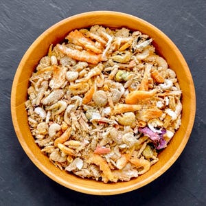 May include: A round wooden bowl filled with a mixture of dried small shrimp, various grains, and small pieces of dried vegetables. The contents feature shades of orange, tan, beige, and green, creating a textured, granular appearance.