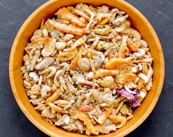 May include: A round wooden bowl filled with a mixture of dried small shrimp, various grains, and small pieces of dried vegetables. The contents feature shades of orange, tan, beige, and green, creating a textured, granular appearance.