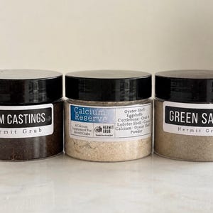 May include: Three clear plastic jars with black lids containing hermit crab food. The labels read Worm Castings, Calcium Reserve with ingredients like Oyster Shell and Cuttlebone, and Green Sand. Each jar is labeled with the brand name Hermit Grub.