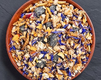 May include: A round wooden bowl filled with a dry mixture of tan mealworms, beige oat flakes, small dark grey charcoal-like nuggets, and bright blue dried cornflower petals. The texture is coarse and varied, showing a blend of organic ingredients.