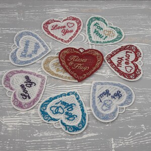 Pode incluir: Nove cora&ccedil;&otilde;es de feltro bordados com diferentes mensagens, incluindo "Love You", "Kisses & Hugs" e "For You!" Os cora&ccedil;&otilde;es est&atilde;o dispostos em c&iacute;rculo sobre uma superf&iacute;cie de madeira cinza.