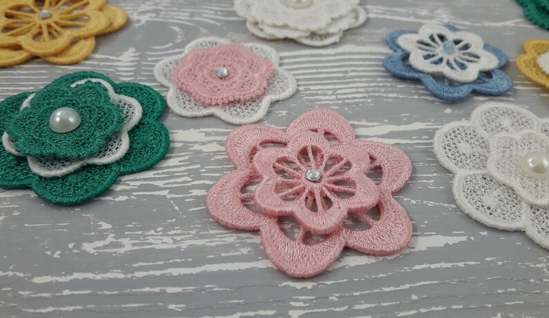 May include: Assortment of layered fabric flower embellishments with pearl and rhinestone accents. The flowers are in shades of pink, green, blue, white, and yellow.