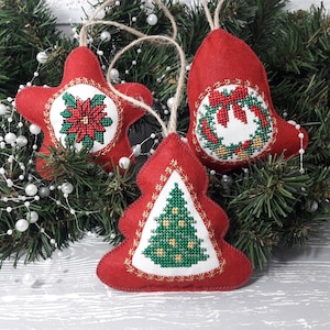 Puede incluir: Tres adornos navideños de fieltro rojo con diseños bordados. Los adornos son una estrella con una flor de pascua, una campana con una corona y un árbol de Navidad.