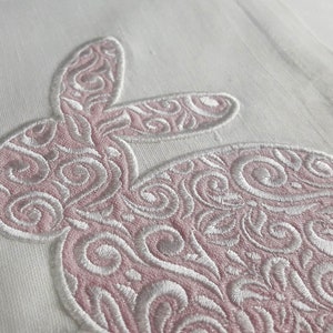 May include: Close-up of a white fabric with a pink embroidered bunny design. The bunny is filled with intricate swirling patterns and outlined in white thread. The design is likely for Easter or spring decor.