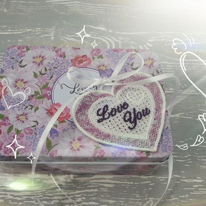 Pode incluir: Uma caixa de presente branca com um padr&atilde;o floral e uma fita branca amarrada ao redor. A fita est&aacute; amarrada em um la&ccedil;o e tem um remendo em forma de cora&ccedil;&atilde;o rosa com as palavras "Love You" bordadas.