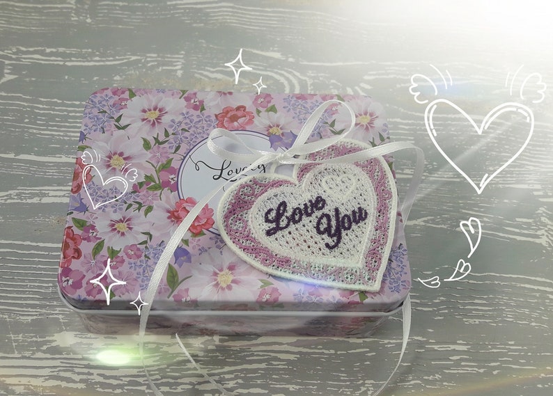 Pode incluir: Uma caixa de presente branca com um padr&atilde;o floral e uma fita branca amarrada ao redor. A fita est&aacute; amarrada em um la&ccedil;o e tem um remendo em forma de cora&ccedil;&atilde;o rosa com as palavras "Love You" bordadas.